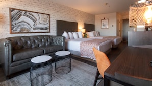 A hotel room with a leather sofa, two round tables, a bed with white bedding, a large abstract painting, and a modern chandelier.