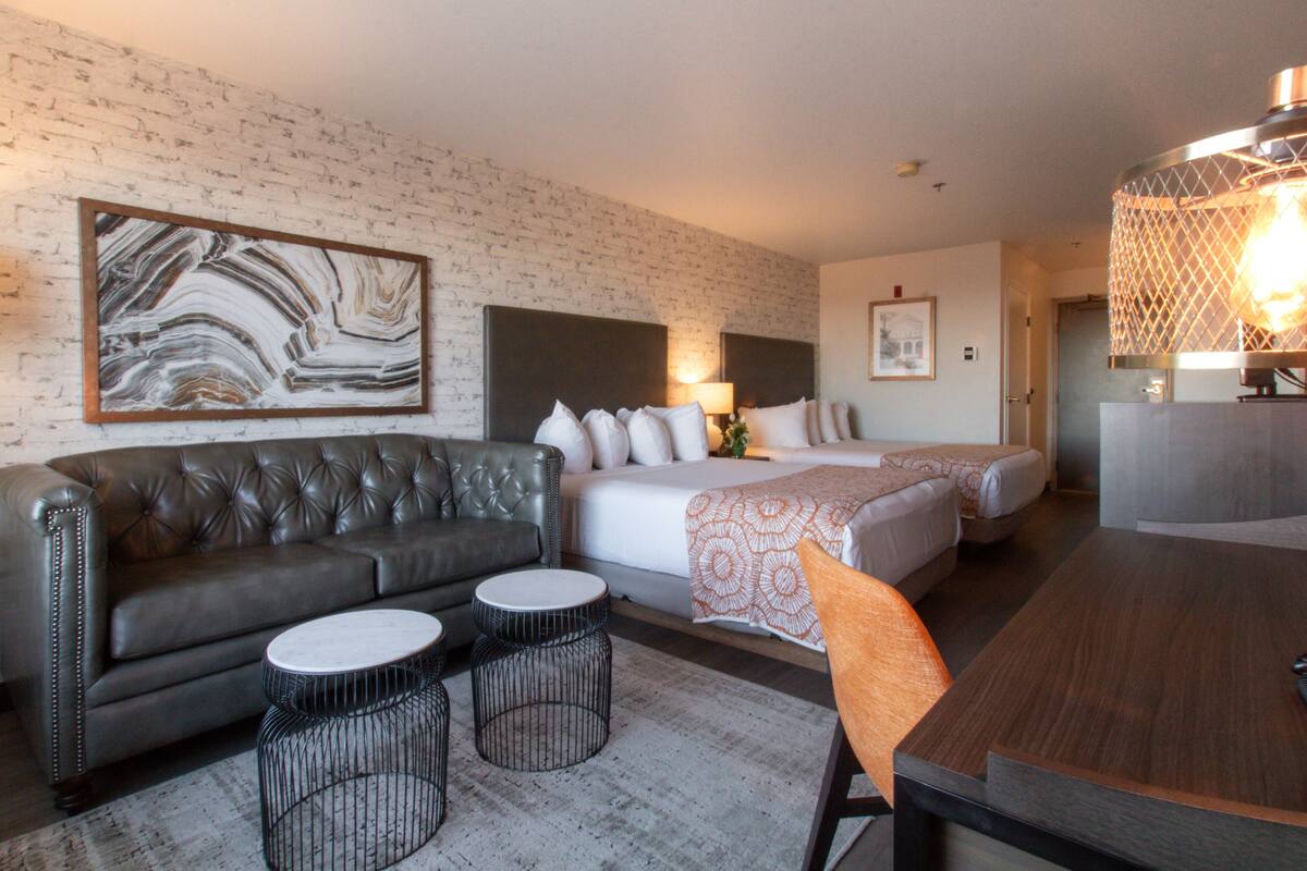 Une chambre d’hôtel avec un canapé en cuir, deux tables rondes, un lit avec une literie blanche, un grand tableau abstrait et un lustre moderne.