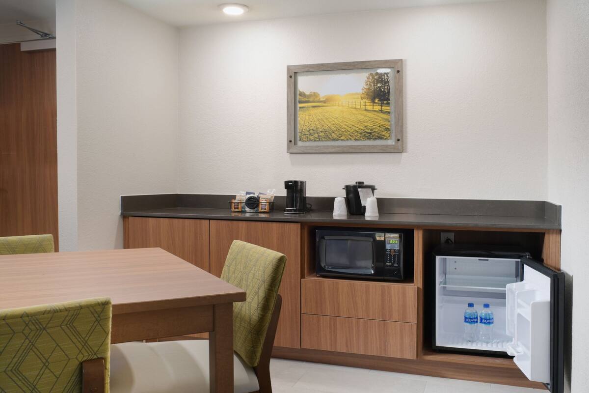 A compact kitchen with a microwave, coffee maker, and a refrigerator. There is a dining table with chairs and a framed picture on the wall.