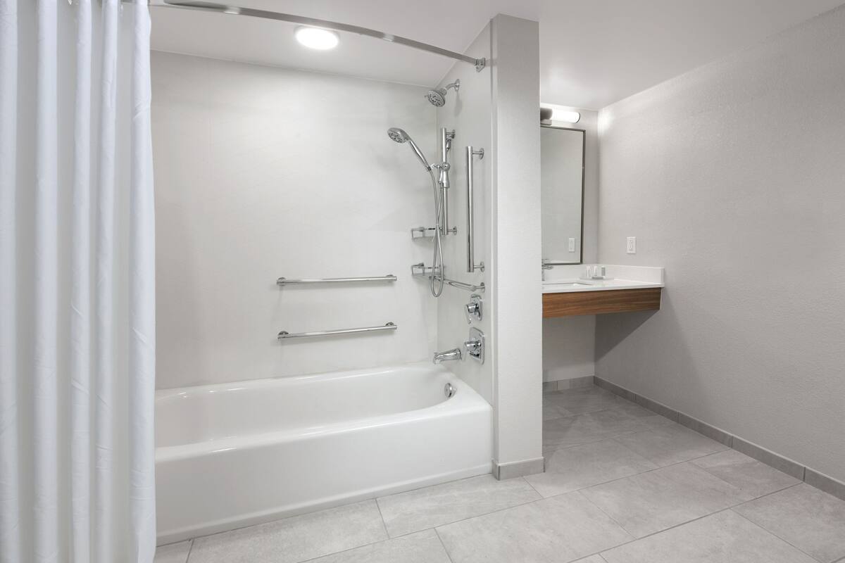 A modern bathroom with a white bathtub, a shower with multiple heads, and a wooden vanity with a mirror.