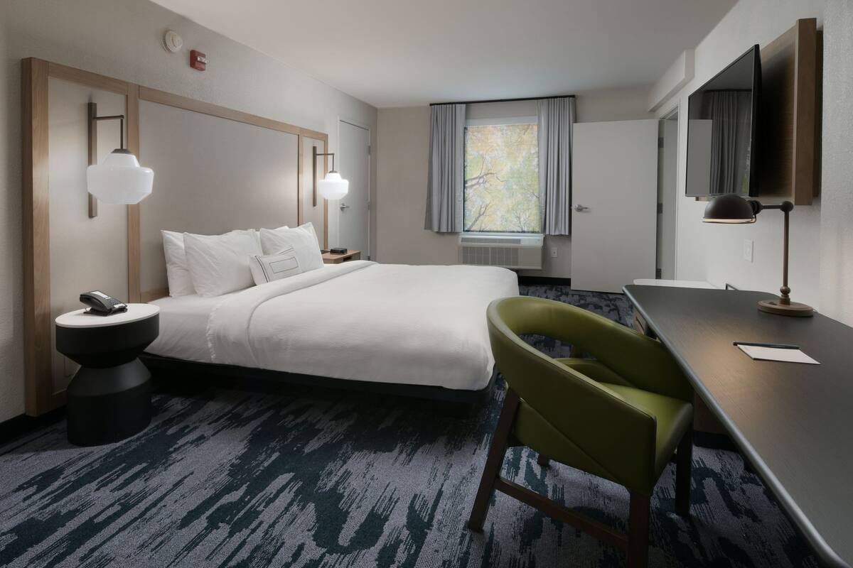 A hotel room with a bed, bedside table, lamp, chair, desk, and window.