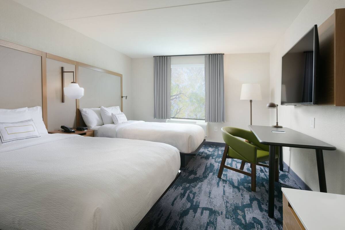 A hotel room with two beds, a desk, a chair, a TV, and a window with curtains.