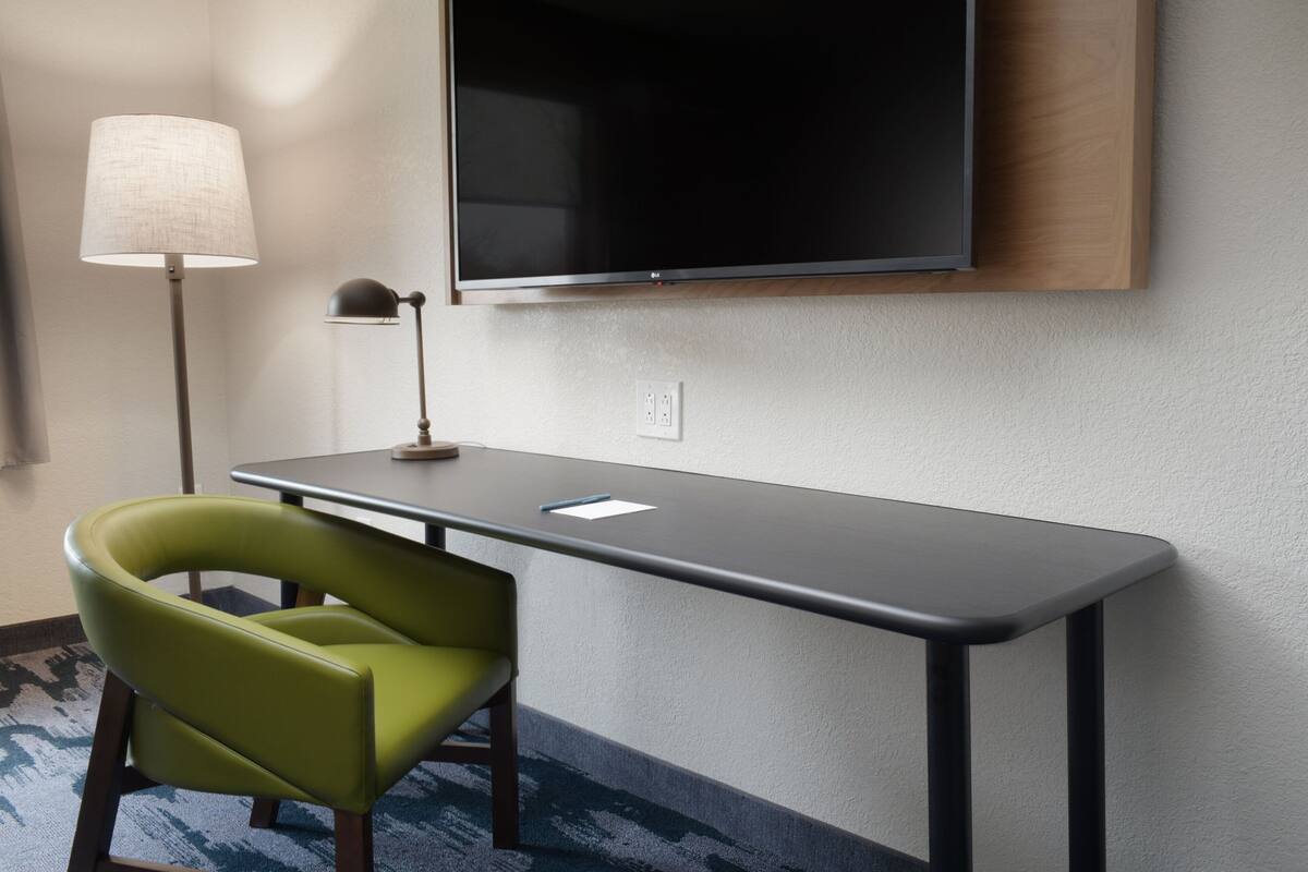 A hotel room with a desk, a green chair, a lamp, and a flat-screen TV mounted on the wall.