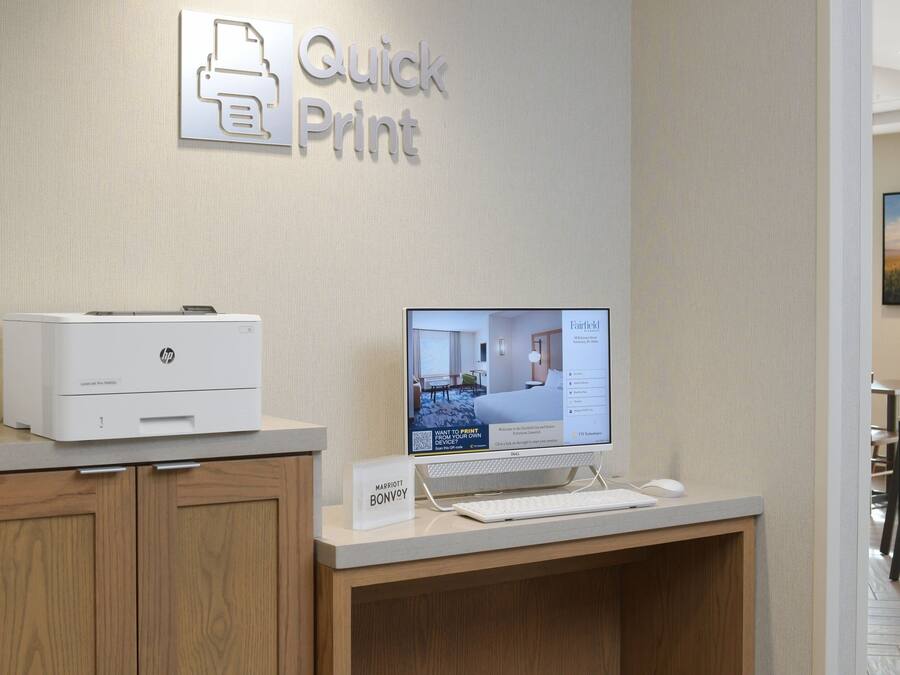 Een receptiebalie in een hotel, met een HP-printer, een computerbeeldscherm waarop een uitzicht op een kamer te zien is, en een bord met de hotelnaam.