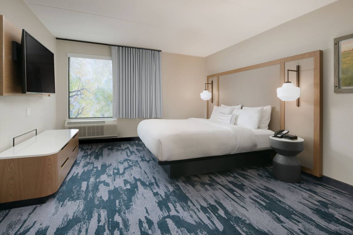 A hotel room with a bed, bedside table, lamp, and a large window with curtains.