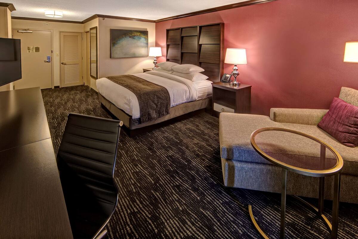 A hotel room with a bed, a desk, a chair, a small table, and a TV.