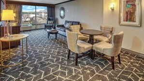 A hotel room with a large window, a sofa, a dining table with chairs, and a lamp.