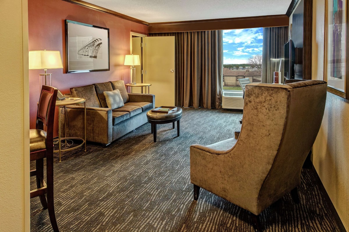 A hotel room with a sofa, armchair, and a small table. There is a window with curtains, a framed picture on the wall, and a lamp on the side table.