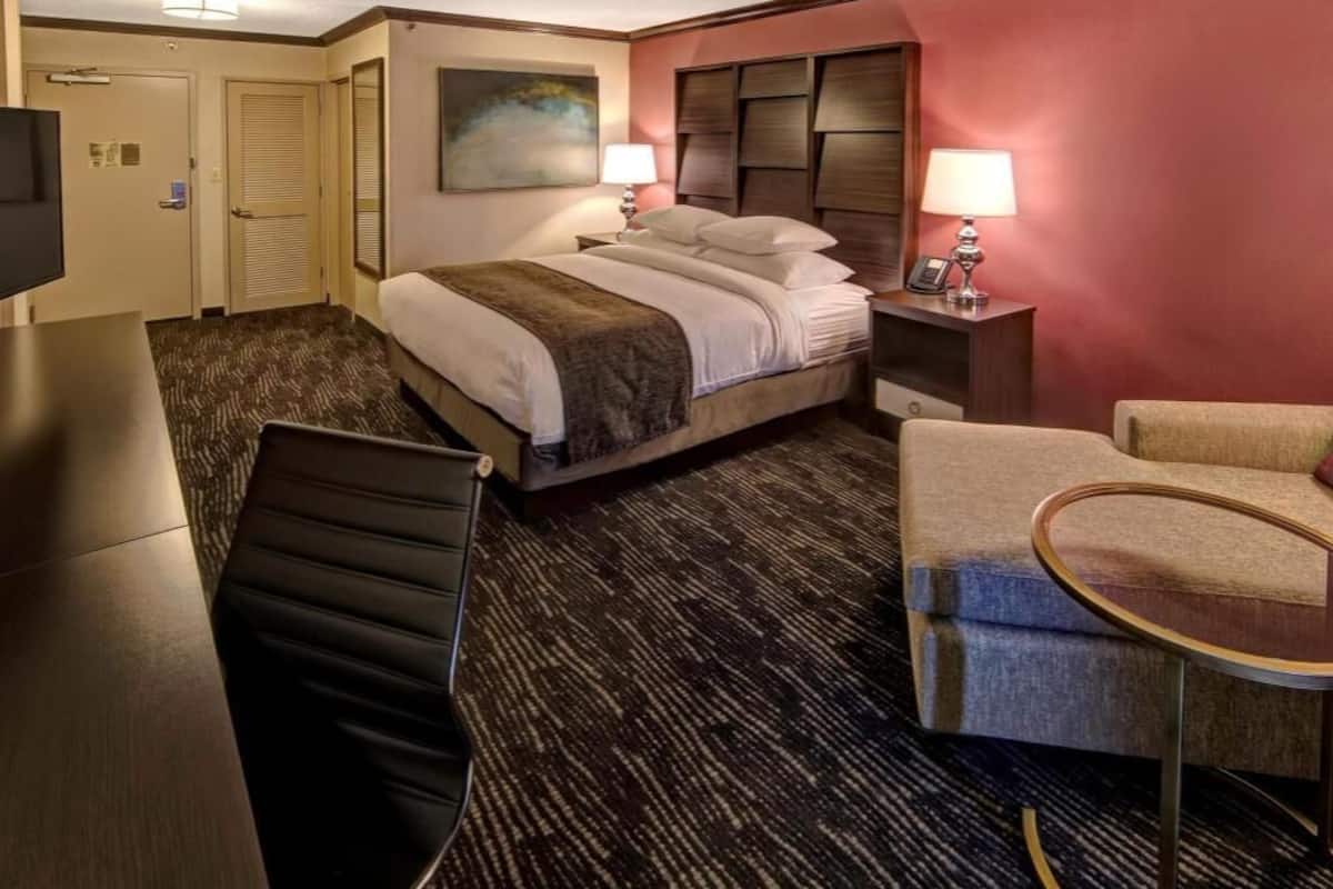 A hotel room with a bed, a desk, a chair, a small table, and a lamp.