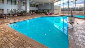 An indoor swimming pool with a tiled deck, lounge area with chairs, and large windows offering a view of the outdoors.