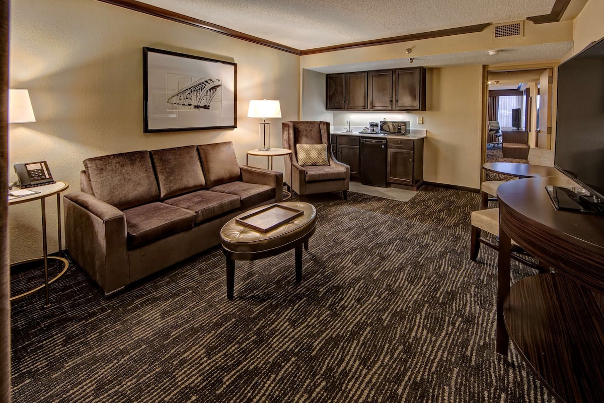 A hotel room with a sofa, armchair, coffee table, kitchenette, and a TV.