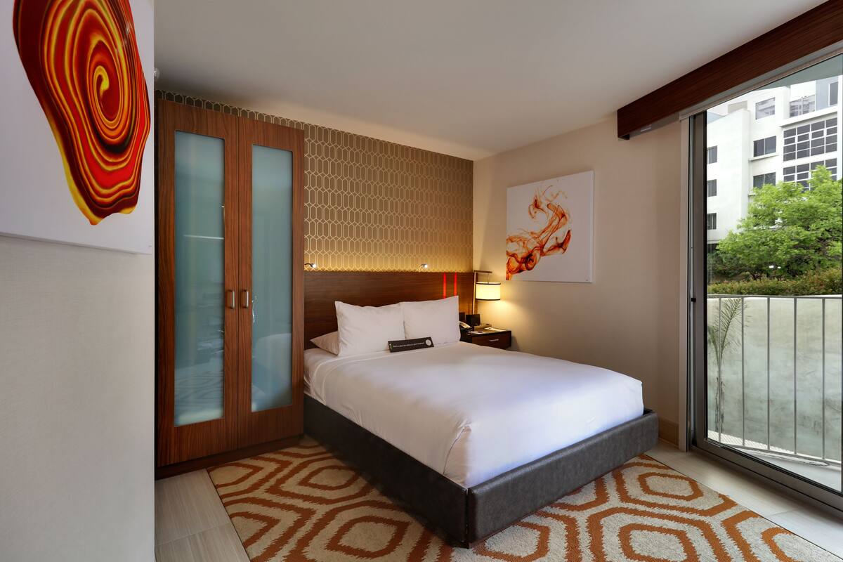 A modern hotel room with a large bed, a wooden wardrobe with glass doors, a patterned rug, and a view of the city through a large window.