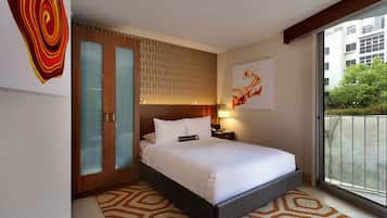 A modern hotel room with a large bed, a wooden wardrobe with glass doors, a patterned rug, and a view of the city through a large window.