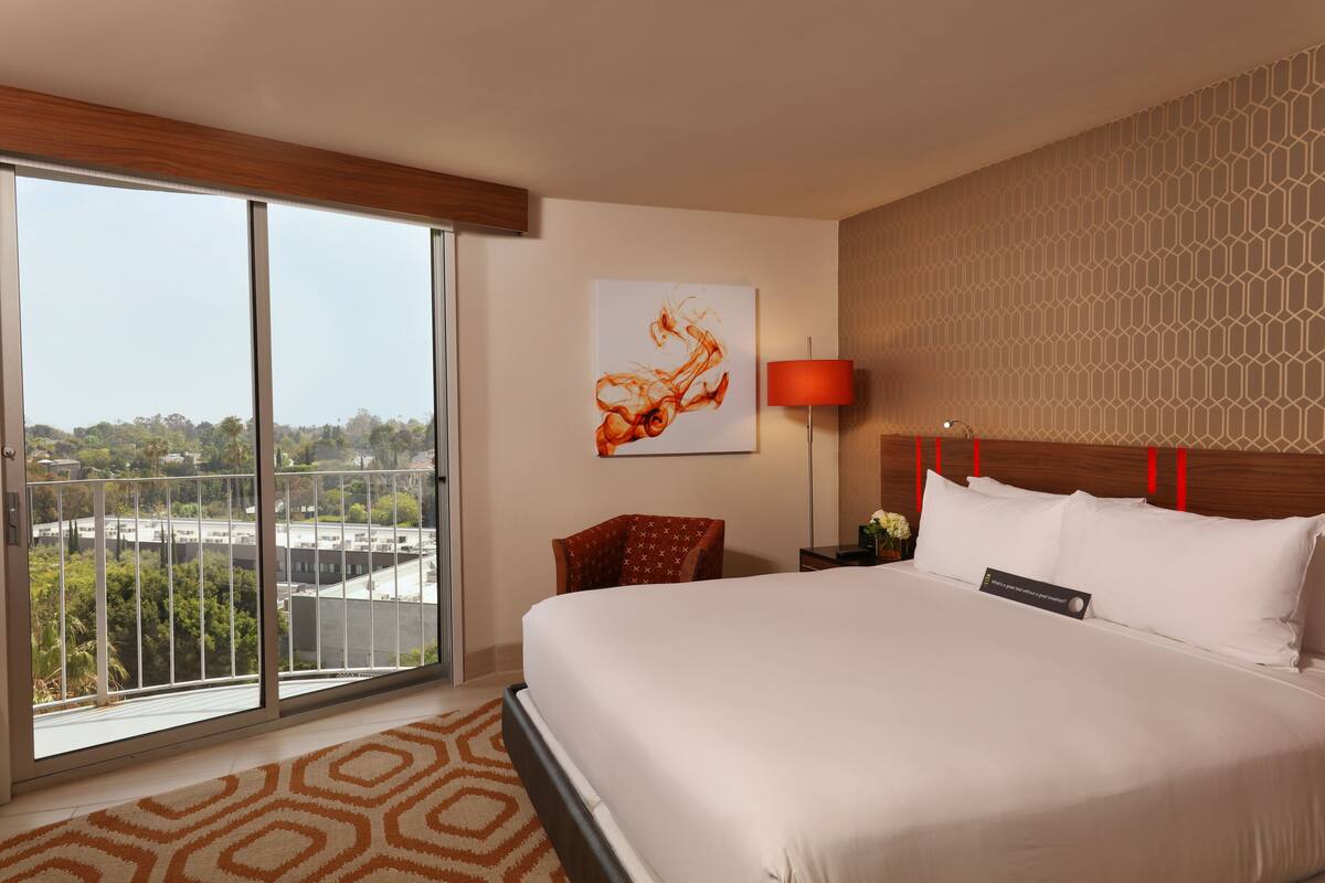 A hotel room with a large bed, a chair, a nightstand, a lamp, and a painting on the wall.