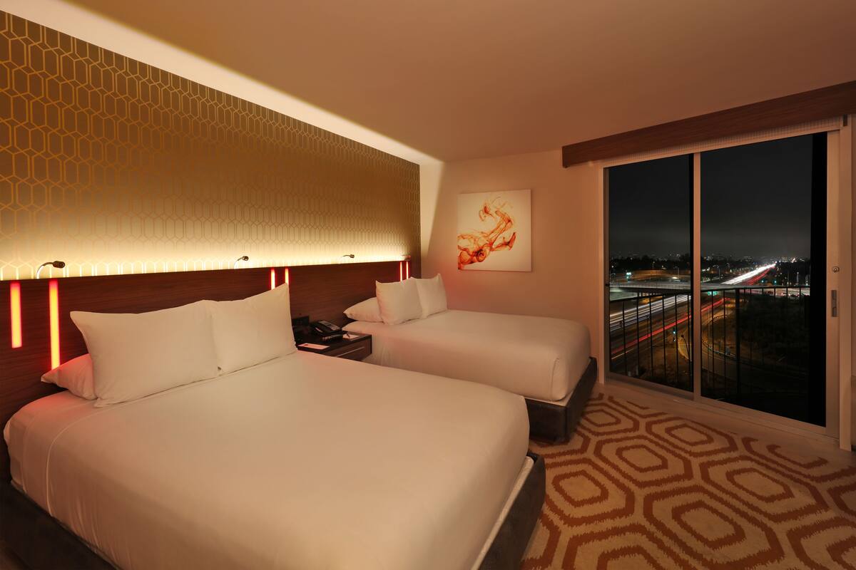 A hotel room with two beds, a large window with a city view, and a patterned carpet.