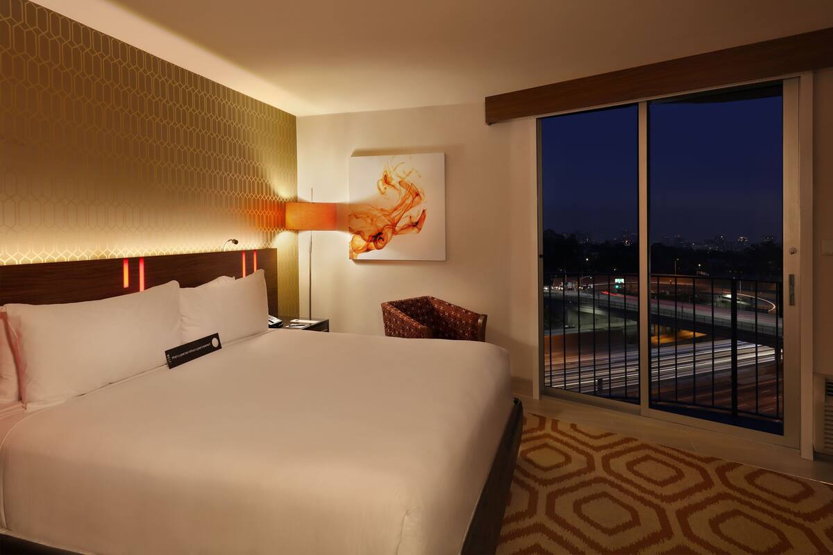 A hotel room with a large bed, a nightstand, a lamp, a chair, and a large window with a city view.