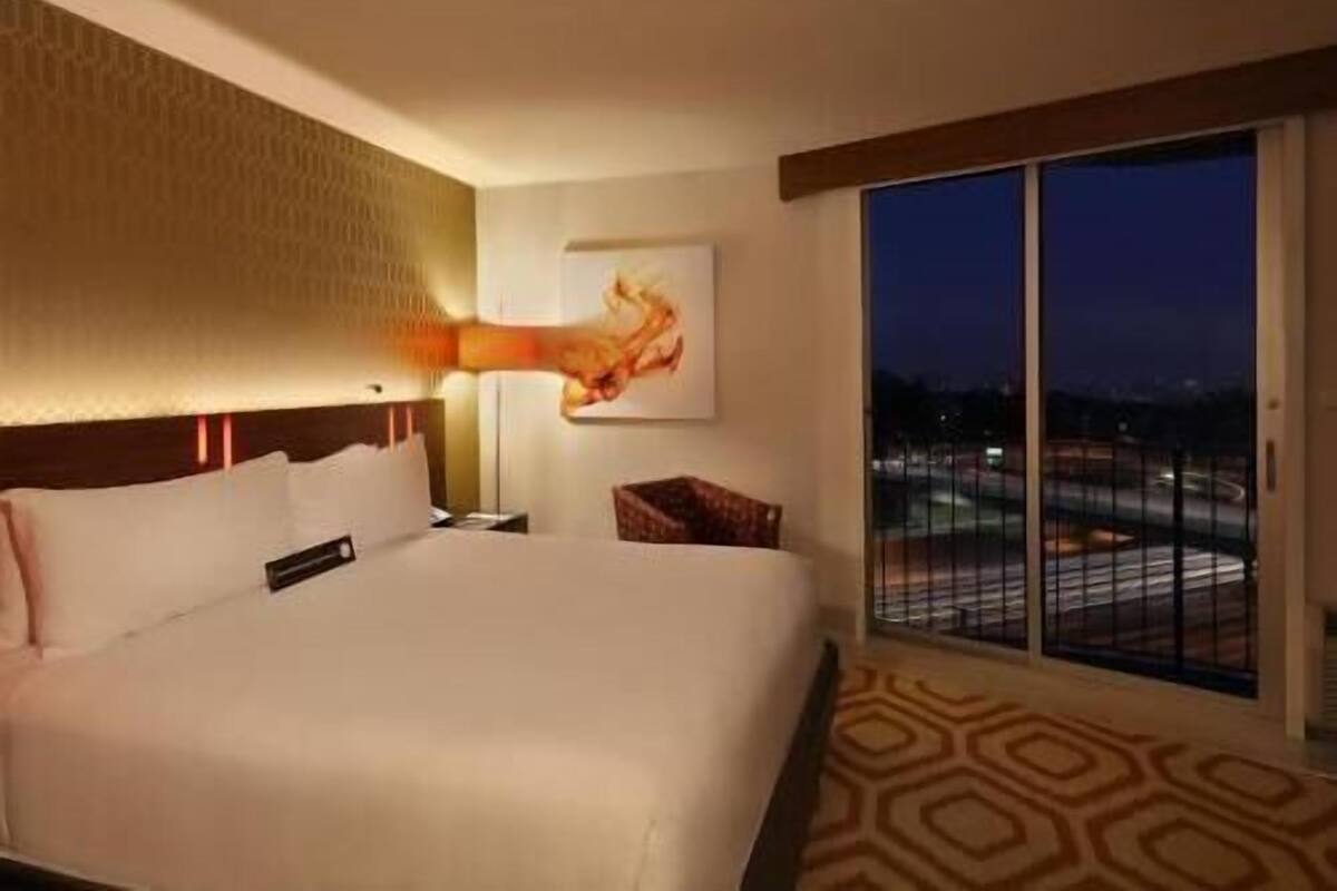 A hotel room with a large bed, a nightstand, a lamp, a painting, and a view of the city at night.