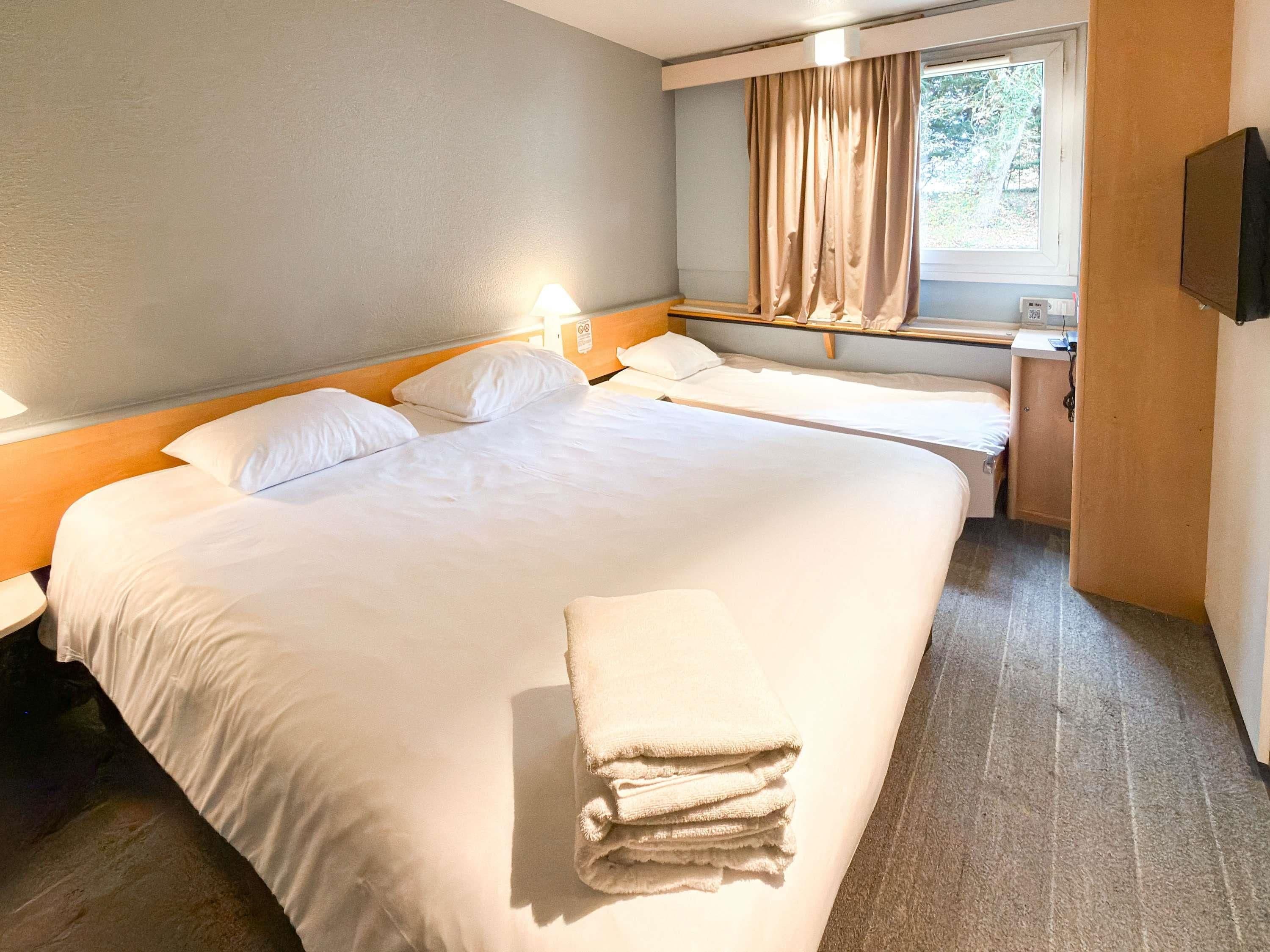 Quarto de hotel com uma cama grande, duas camas menores, uma escrivaninha, uma cadeira, uma televisão e uma janela com cortinas.