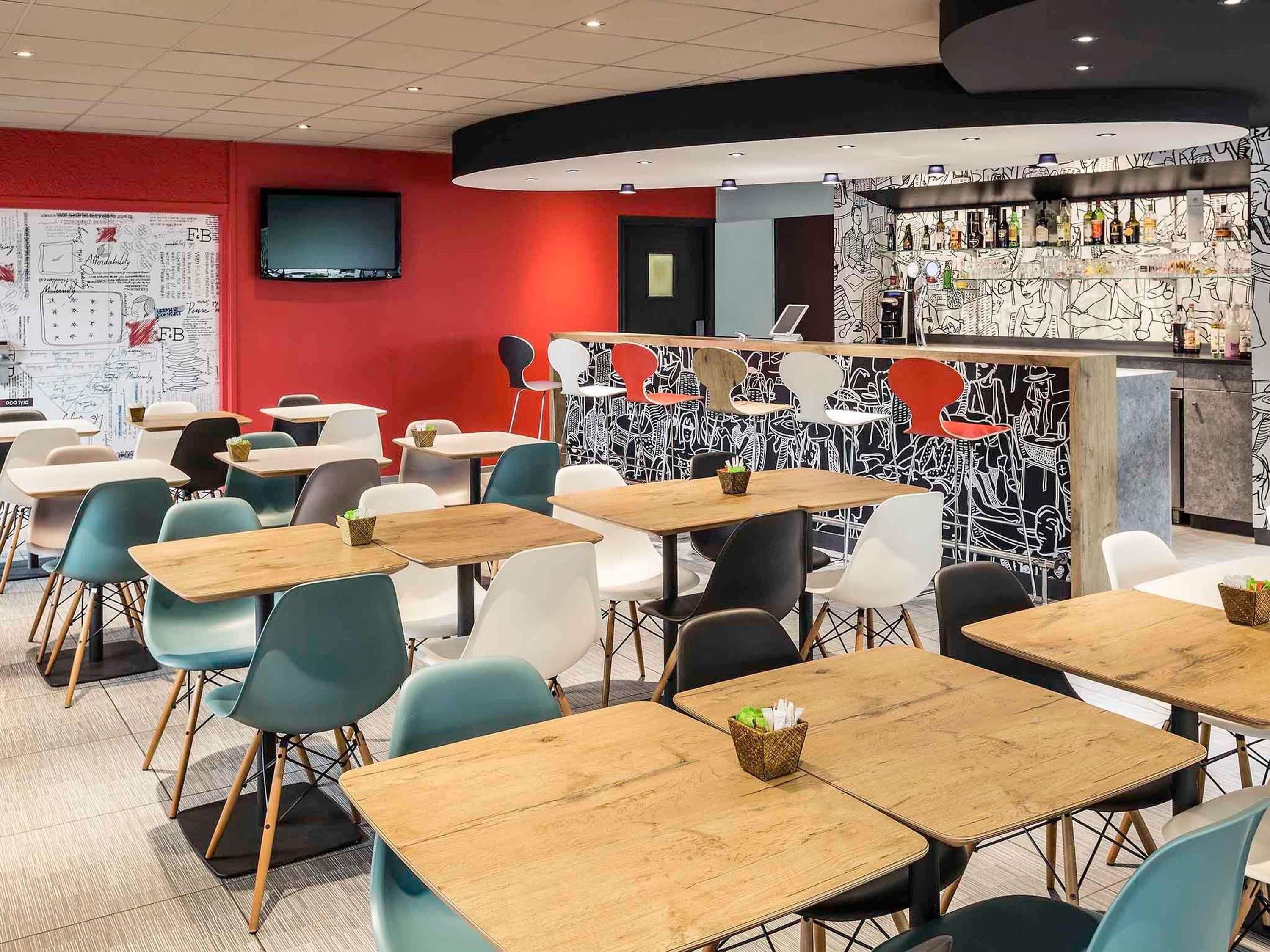 Un café moderne doté de nombreuses tables et chaises, d’un bar agrémenté d’une fresque murale originale en noir et blanc, et d’un téléviseur à écran plat fixé au mur.
