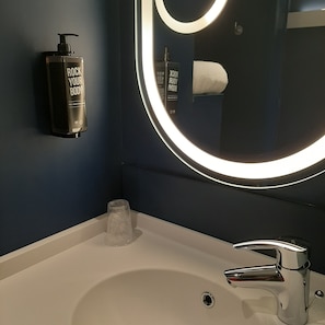 Un baño con un lavamanos blanco, un espejo con luz circular, una botella de crema corporal y un vaso de agua.