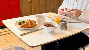 A breakfast table with a croissant, a bowl of cereal, a coffee cup, a glass of orange juice, and a metal strainer. - ibis Rennes Cesson (Cesson-Sevigne)