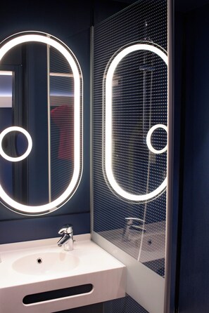 Un baño con un lavamanos blanco, un espejo con luz circular y una zona de ducha.
