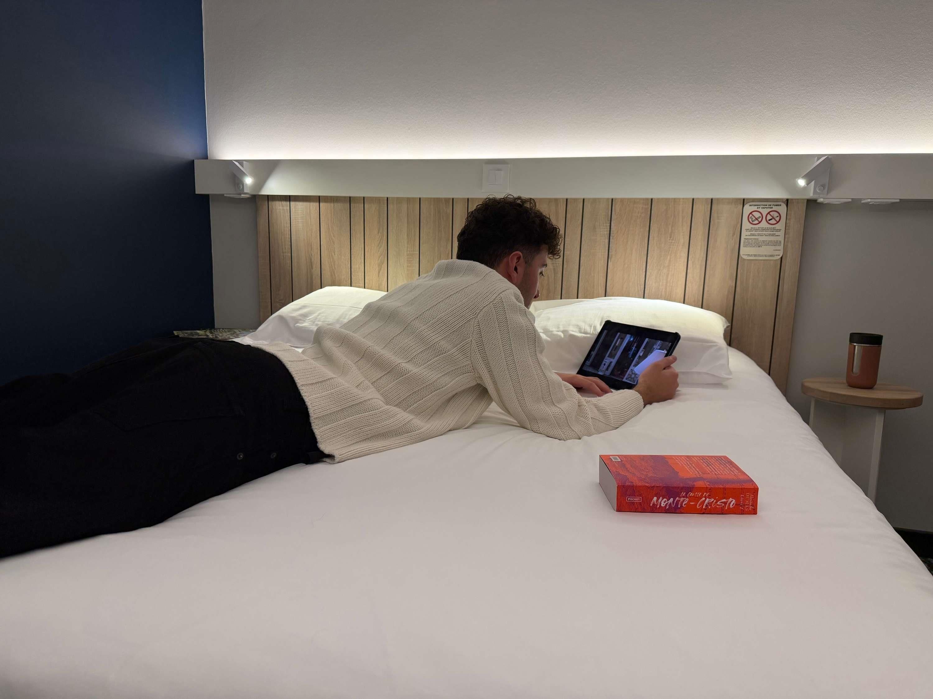 En person ligger i sengen med en tablet, en bog og en kop på et natbord.