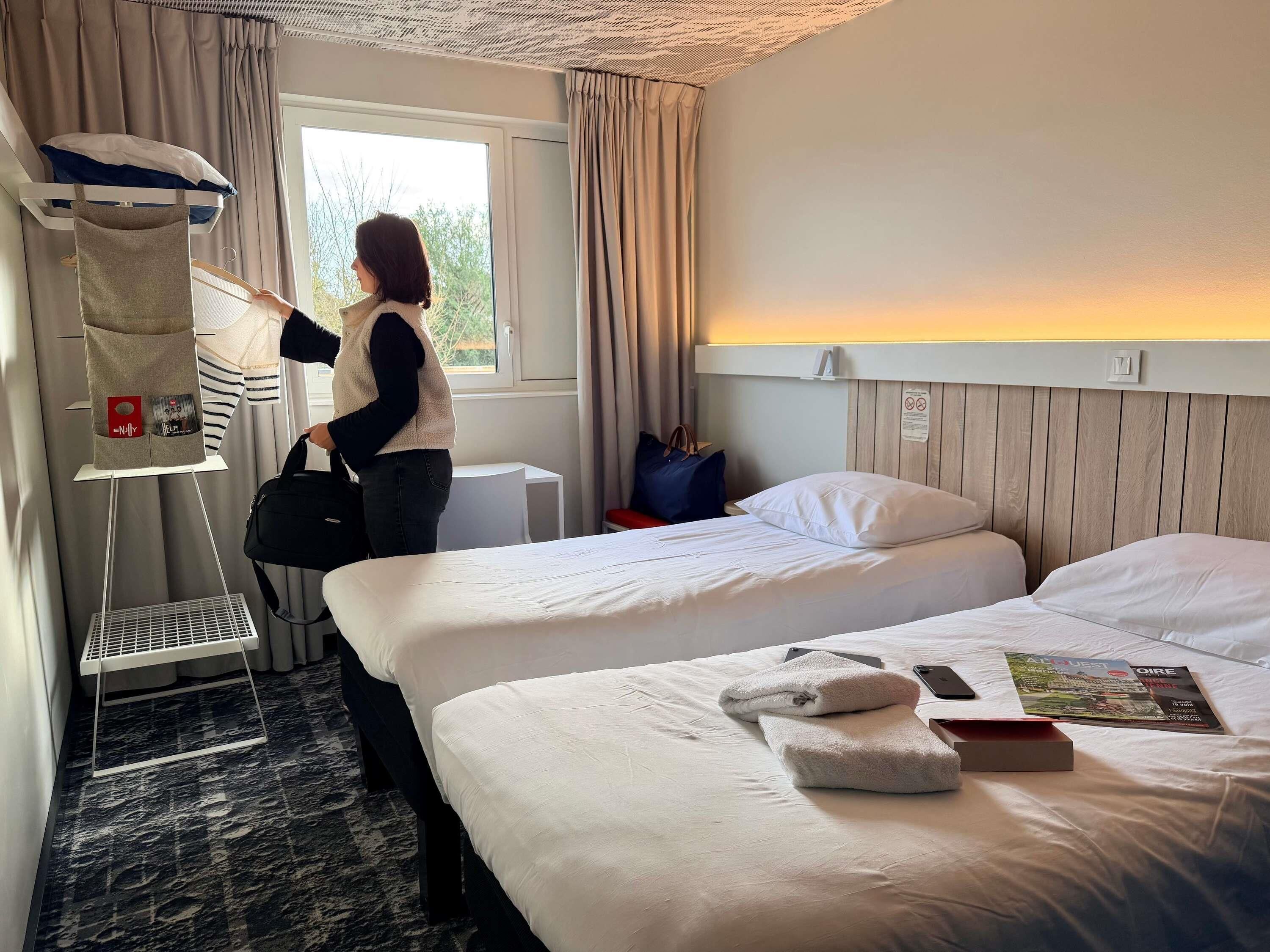 Habitación de hotel con dos camas, una ventana con cortinas, una mesita de noche y un estante en la pared con un bolso.