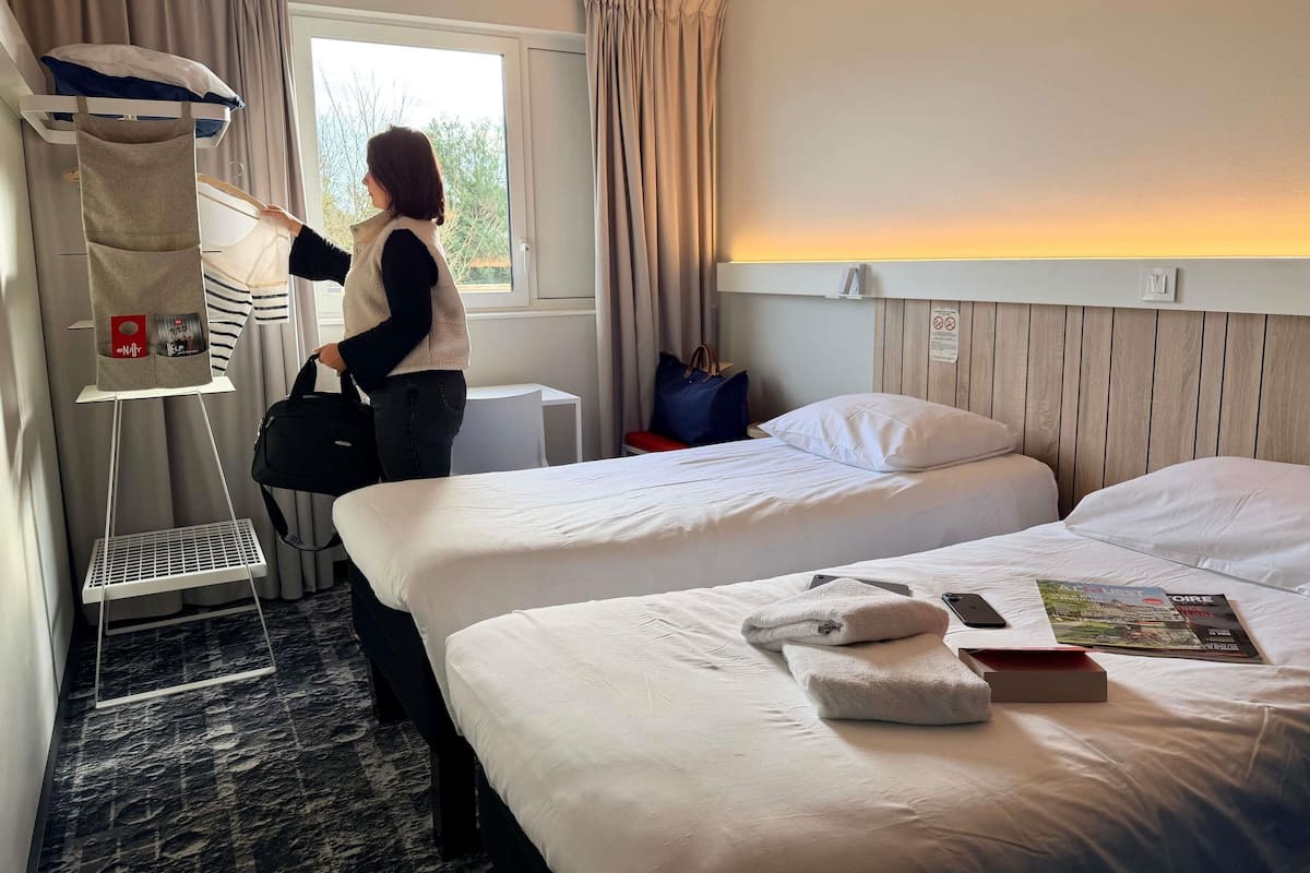 Quarto de hotel com duas camas, uma janela com cortinas, uma mesa de cabeceira e uma prateleira na parede com uma bolsa.