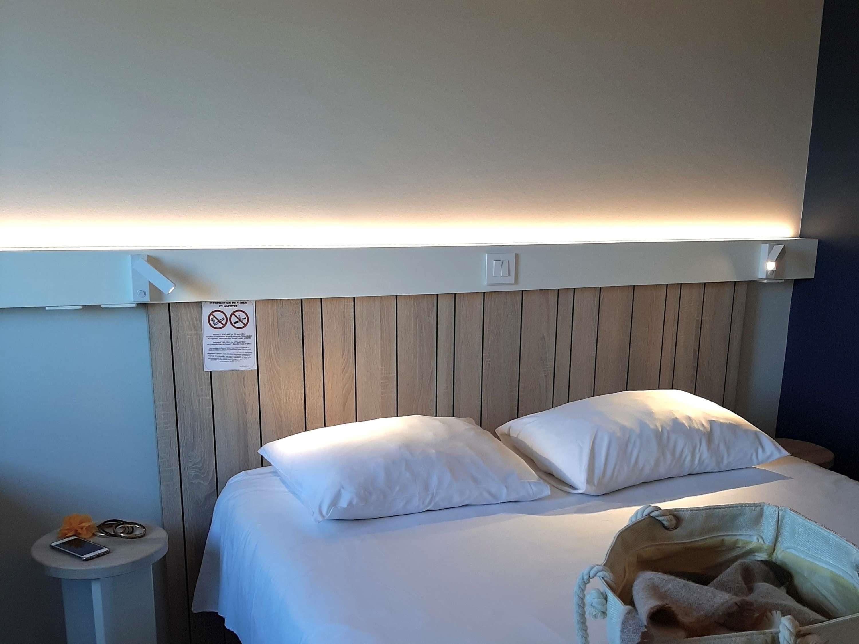 Une chambre d’hôtel avec une tête de lit en bois, deux oreillers blancs, une table de chevet avec un téléphone et des clés, et une petite poubelle.