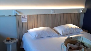 A hotel room with a wooden headboard, two white pillows, a bedside table with a phone and keys, and a small trash bin. - ibis Rennes Cesson (Cesson-Sevigne)