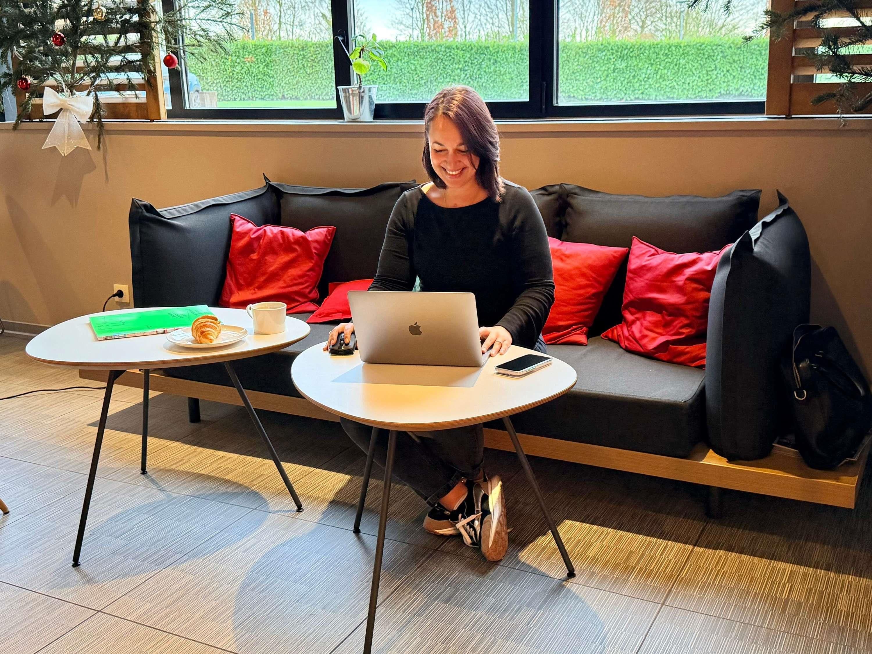 Eine Person sitzt auf einer schwarzen Couch mit roten Kissen und benutzt einen Laptop auf einem kleinen runden Tisch. Auf dem Tisch liegen ein Buch, ein Croissant und eine Tasse.