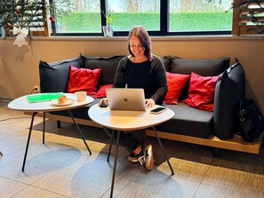 A person sitting on a black sofa with red cushions, using a laptop on a small round table. There's a book, a croissant, and a cup on the table. - ibis Rennes Cesson (Cesson-Sevigne)
