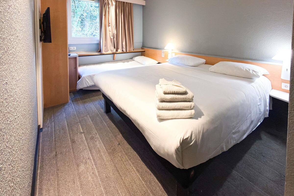 Habitación de hotel con una cama grande, una cama más pequeña, una mesita de noche, una ventana con cortinas y una lámpara fijada a la pared.