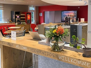 A hotel reception area with a wooden counter, a laptop, a vase with flowers, and a television displaying a scenic view. - ibis Rennes Cesson (Cesson-Sevigne)