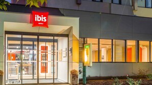 Una entrada de hotel moderna con fachada de vidrio, un letrero rojo que indica que es un hotel ibis y un área de recepción visible.
