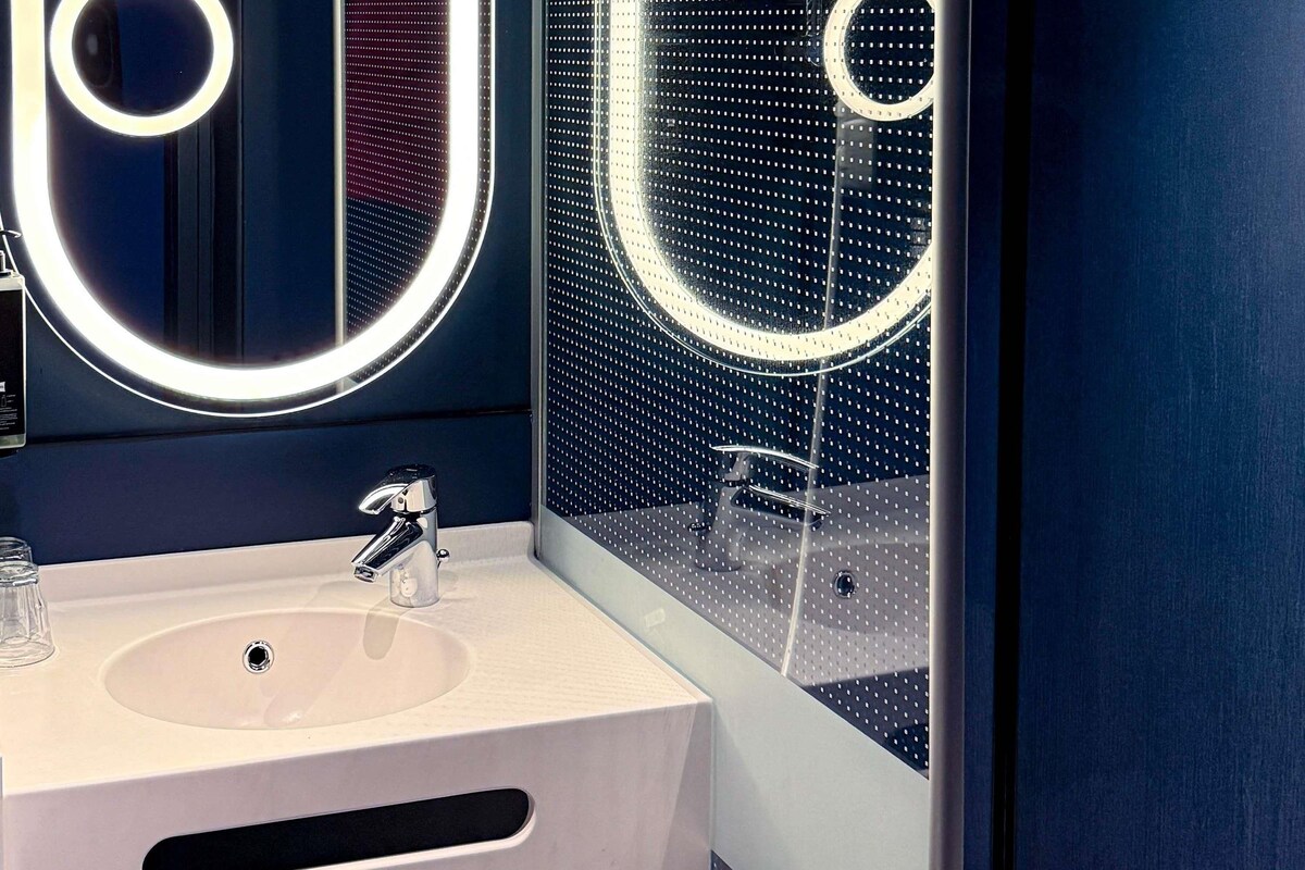 Un baño moderno con un lavamanos blanco, grifo cromado y un espejo grande con luces circulares.