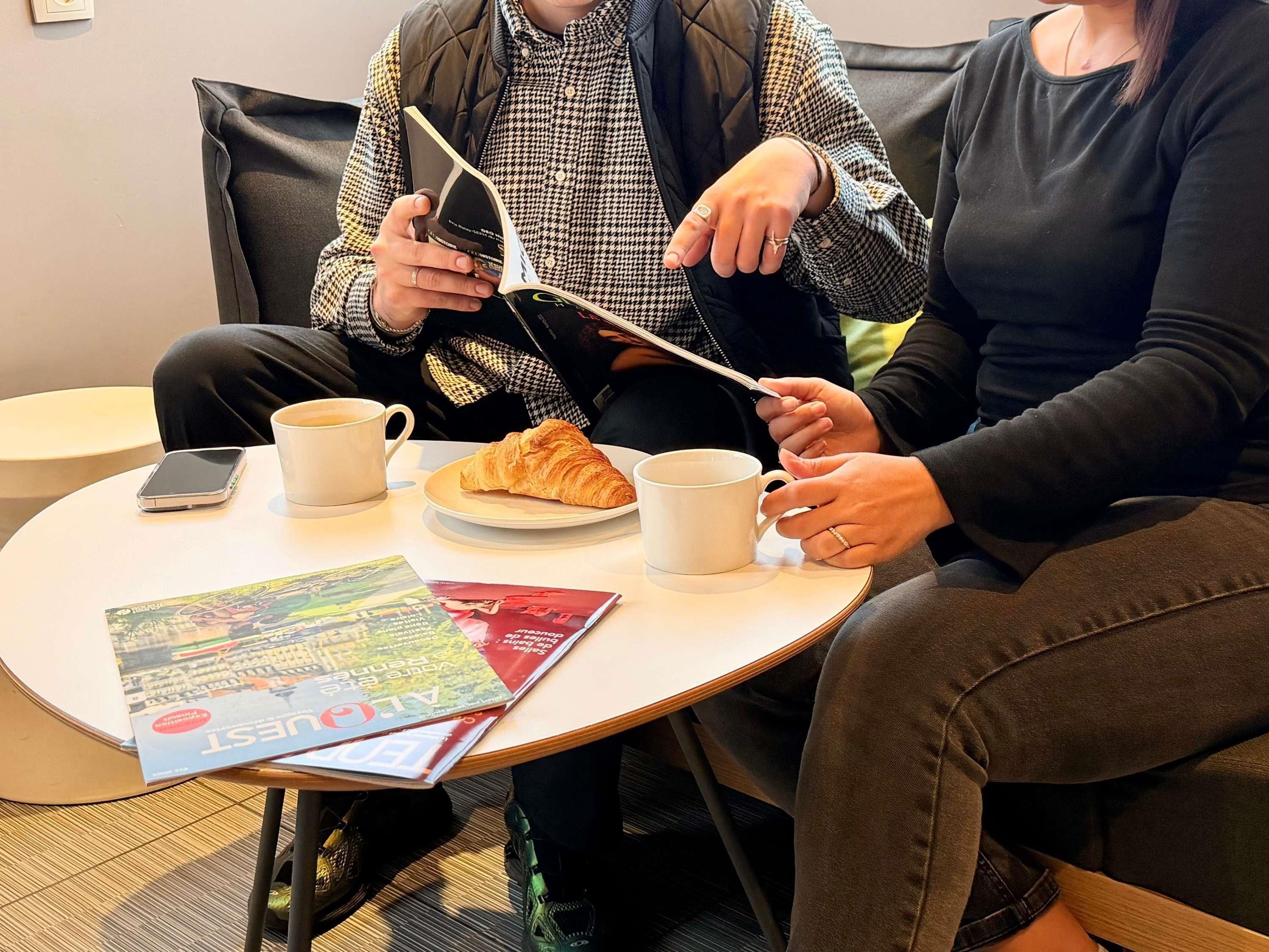 Twee personen zitten aan een ronde tafel met een croissant, twee witte mokken en een smartphone.