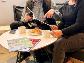 Dos personas sentadas en una mesa redonda con un croissant, dos tazas blancas y un teléfono inteligente.