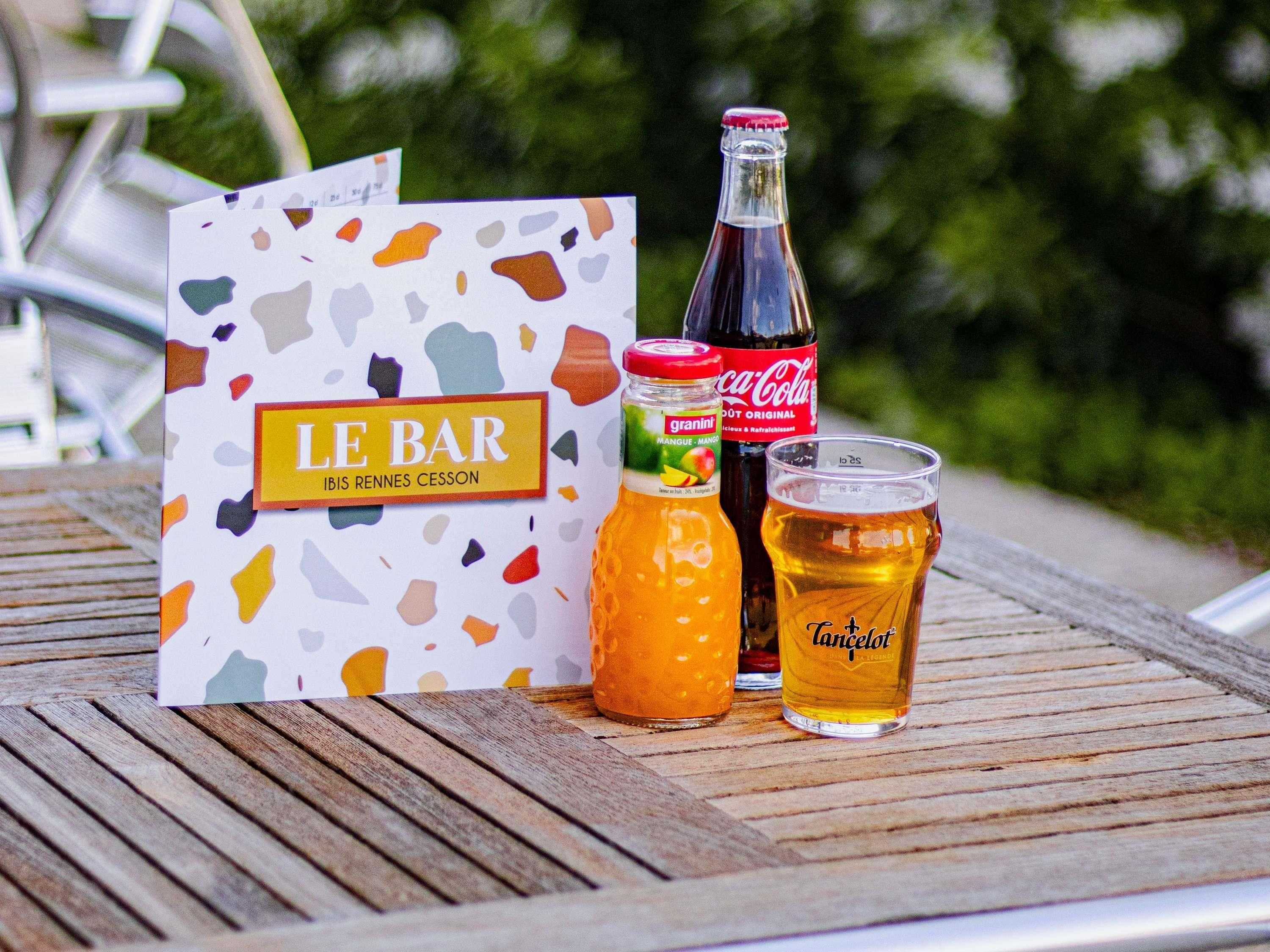 A wooden table with a card labeled 'LE BAR,' a bottle of Granini mango, a Coca-Cola bottle, and a glass of beer.