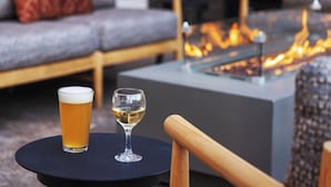 A glass of beer and a glass of white wine on a round table, with a sofa and a fire feature in the background. - Starlite Yellowstone, Tapestry Collection by Hilton (West Yellowstone)