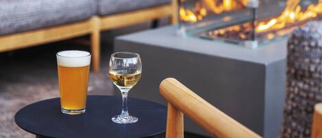 A glass of beer and a glass of white wine on a round table, with a sofa and a fire feature in the background.