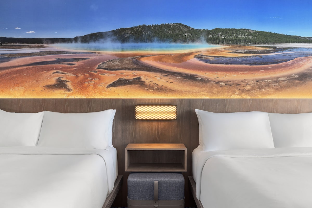 A hotel room with a large window overlooking a geothermal area with steam and colorful mineral deposits. The room includes two beds, a bedside table, and a small storage bench.