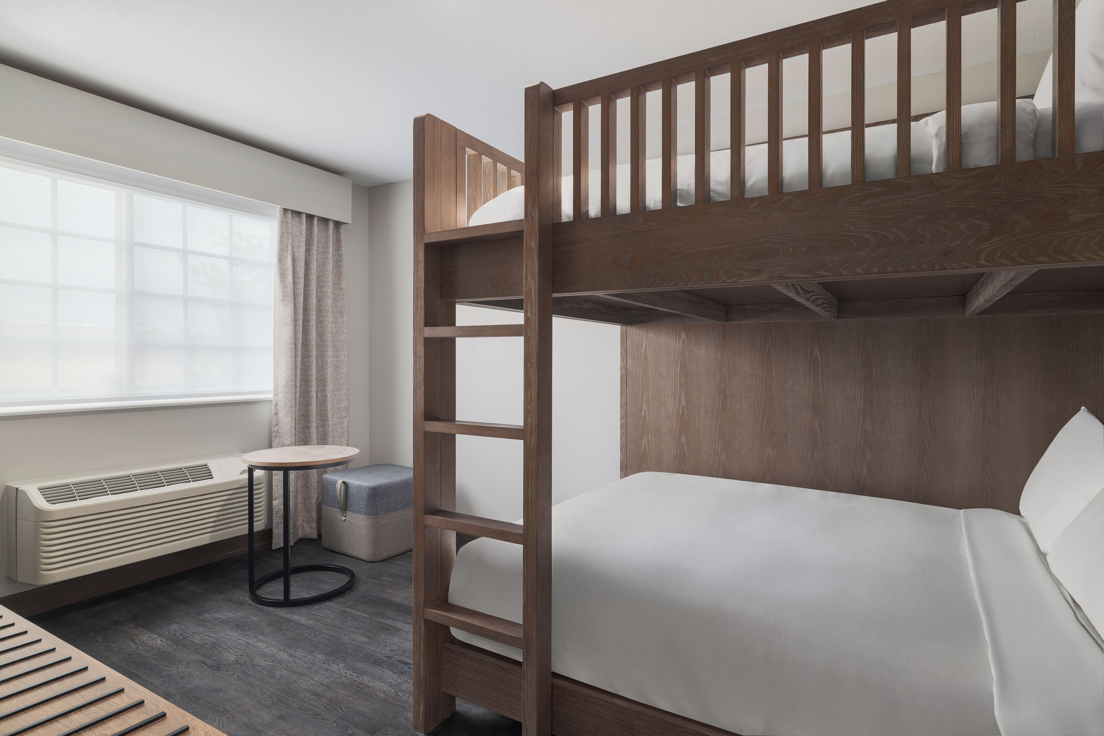 Ein Stockbett mit Holzrahmen und weißer Bettwäsche, ein runder Tisch mit schwarzem Gestell, ein eingebauter Heizkörper und ein Fenster mit Vorhängen.