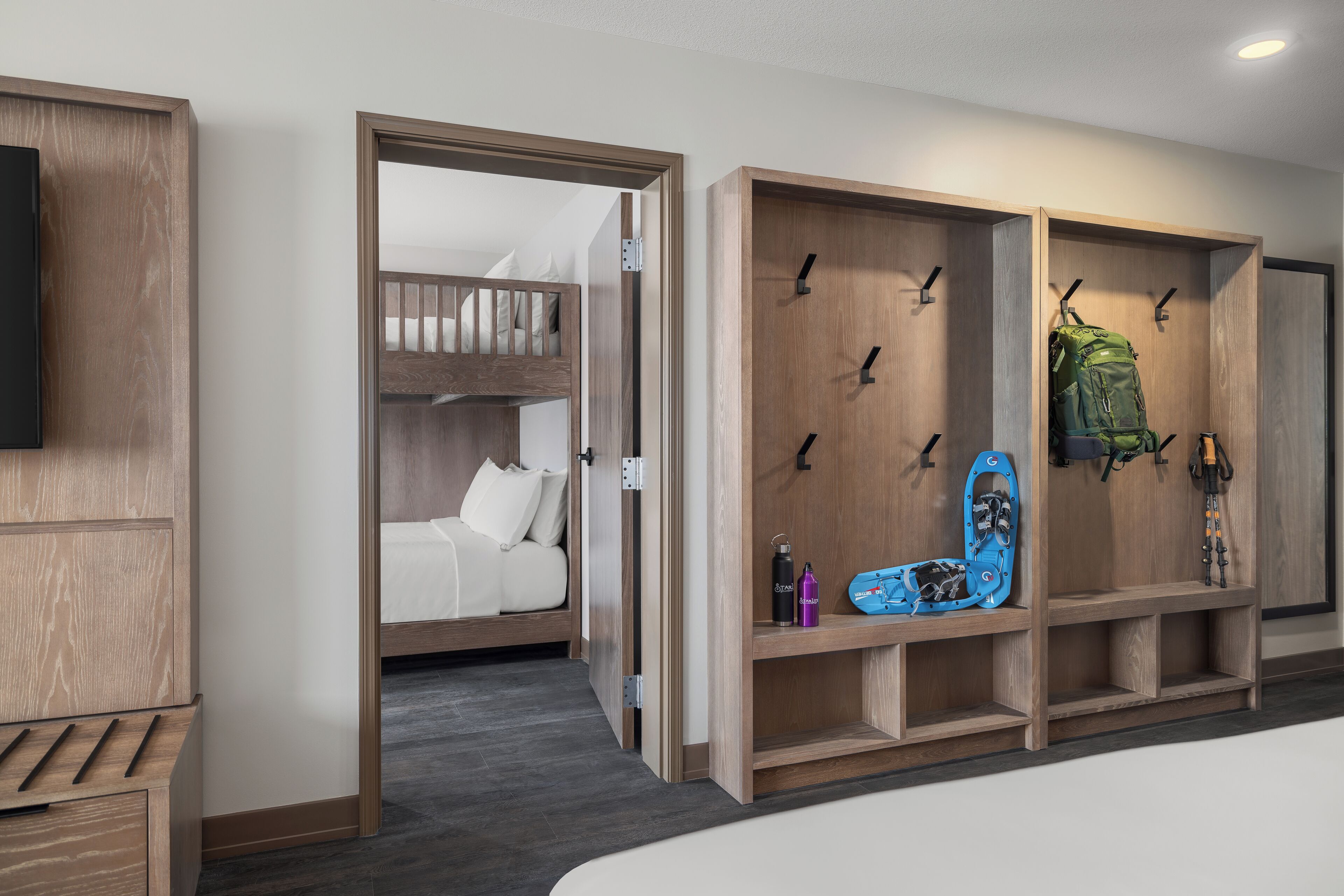Een moderne hotelkamer met een stapelbed, een houten garderobe met haken en een wandgemonteerde televisie.