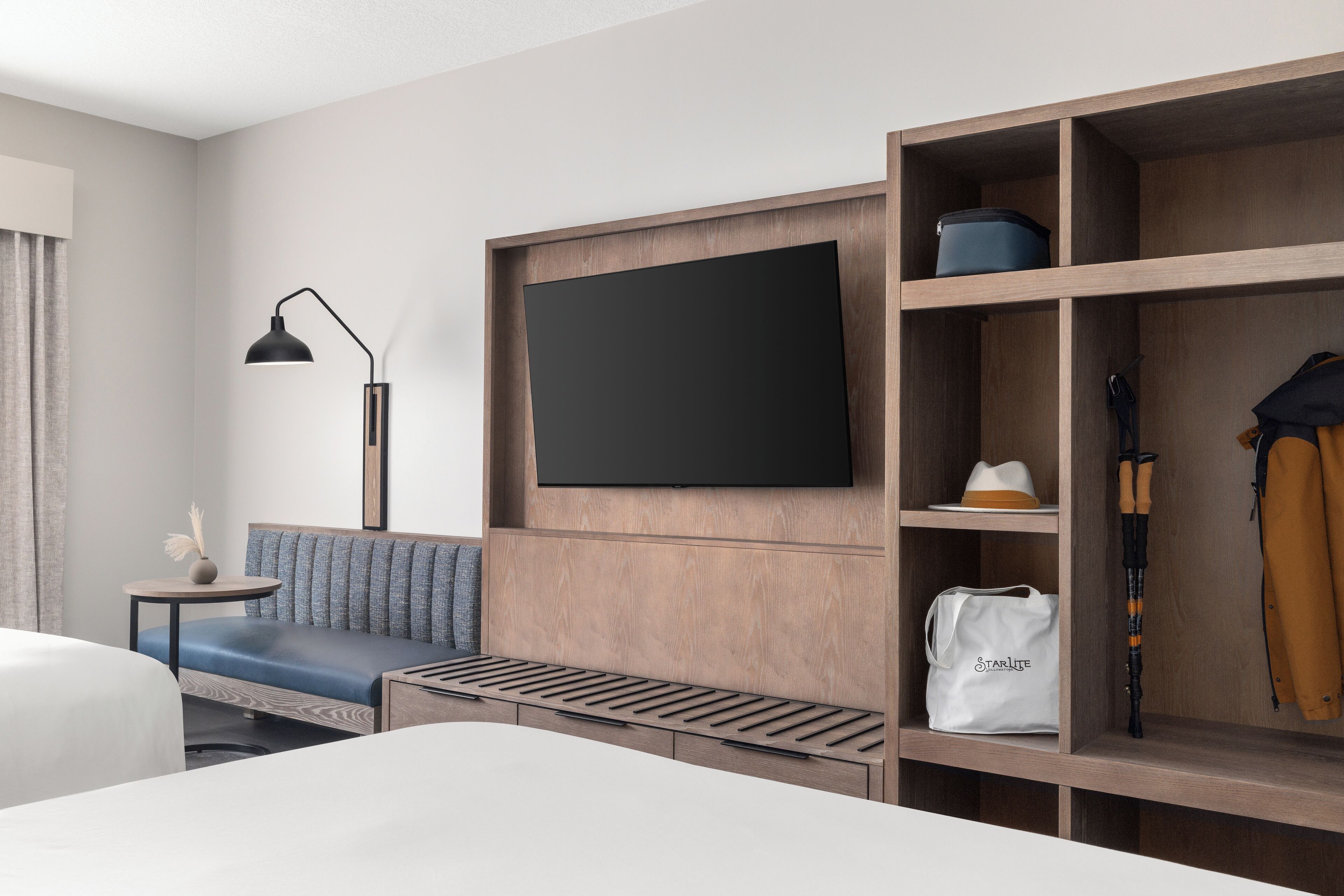 A modern hotel room with a large bed, a sofa, a TV mounted on a wooden panel, a small table with a lamp, and a wardrobe with shelves.