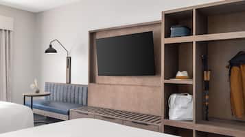 A modern hotel room with a large bed, a sofa, a TV mounted on a wooden panel, a small table with a lamp, and a wardrobe with shelves.