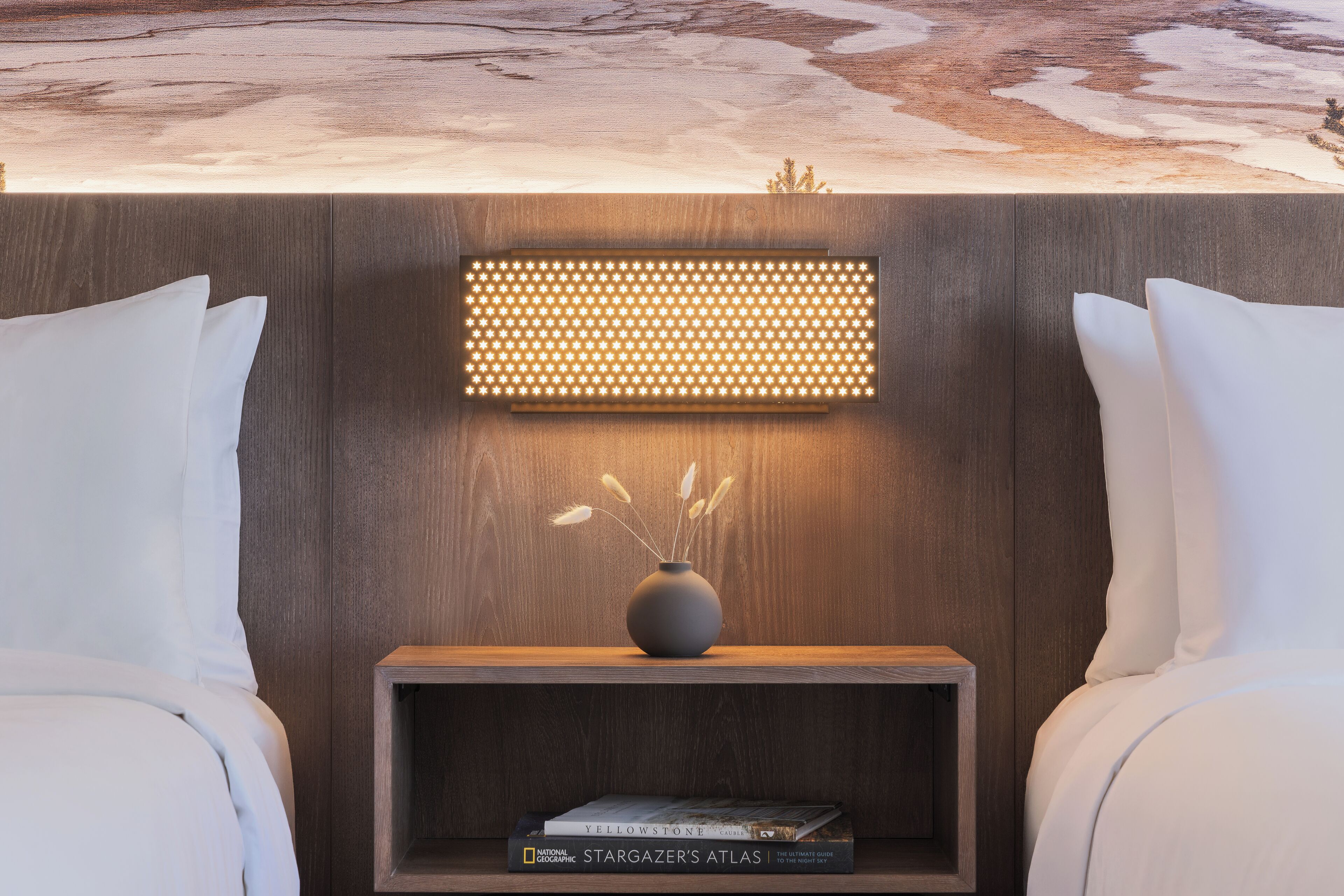 A hotel room with a bed, a bedside table with a vase and books, and a wall-mounted light fixture.