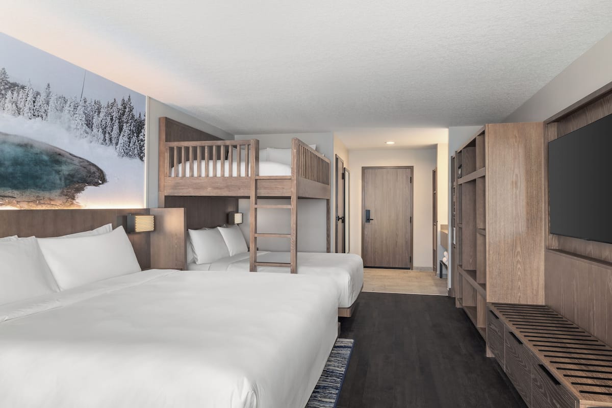 A modern hotel room with a large bed, a bunk bed, a wooden wardrobe, and a painting of a natural landscape.