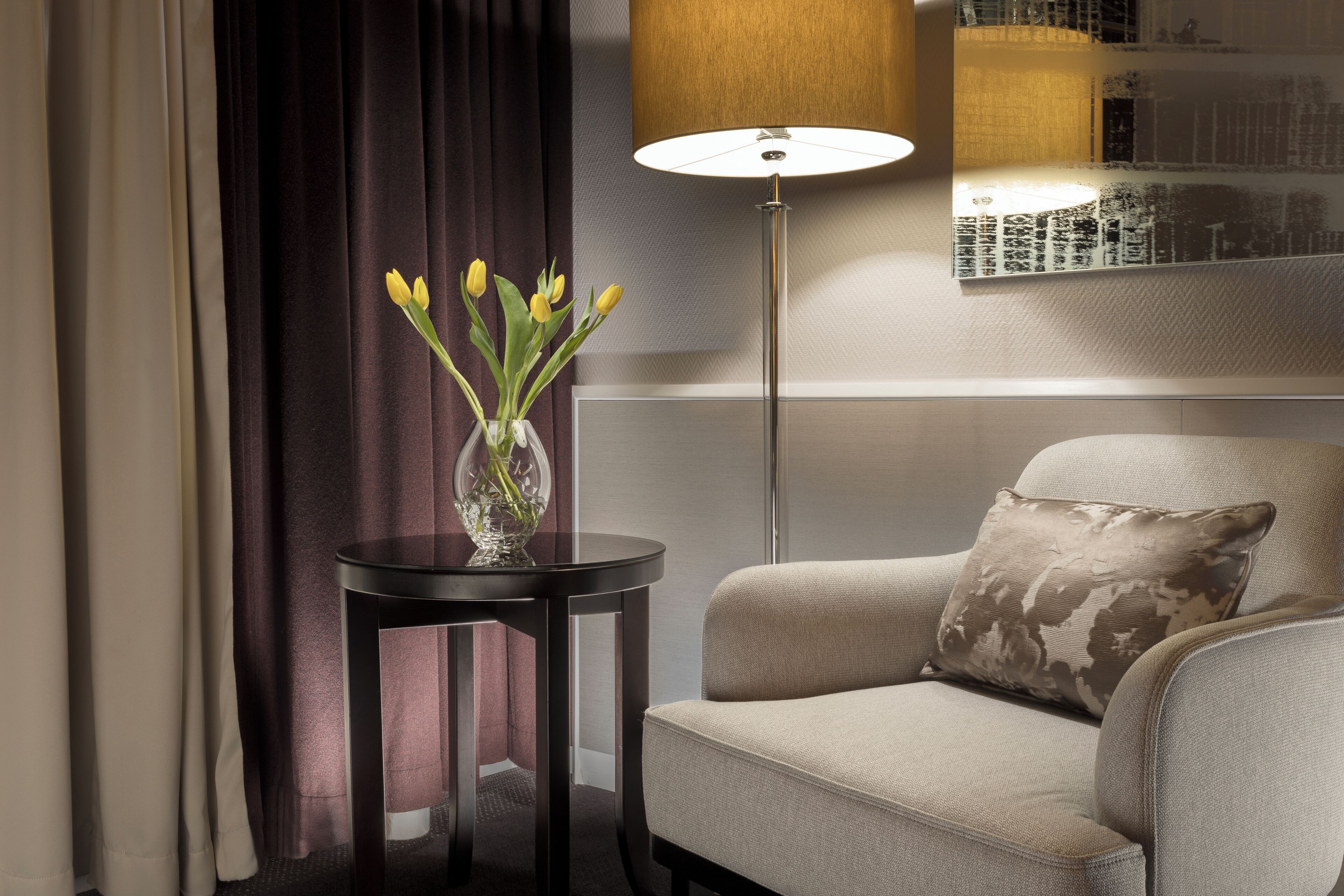A room with a sofa, a small table with a vase of flowers, a floor lamp, and a wall-mounted artwork.